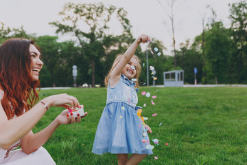 Smiling woman in light dress and little cute child baby girl play with colorful paper confetti in green park. Mother, little kid daughter. Mother's Day, love family, parenthood, childhood concept.