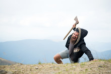 Man With Beard On Shouting Face Conquers Top Of Mountain With Axe, Sky Background. Survive In Wild Nature Concept. Hipster With Axe Surviving In Mountains. Guy Brutal And Bearded In Wild Nature