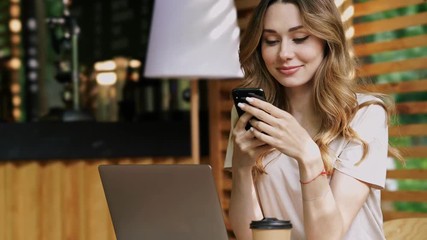 Wall Mural - Close up view of Smiling brunette woman in casual clothes sitting by the table with laptop computer while using smartphone at cafe outdoors