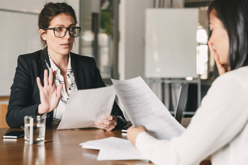 Poster - Image of nervous unsure asian woman talking and negotiating with businesswoman, while sitting at table in office during job interview - business, career and recruitment concept