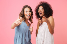 Two Cheerful Elegant Girls 20s With Different Color Of Skin In Dresses Smiling And Pointing Fingers At Camera Meaning Hey You, Isolated Over Pink Background