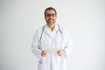 Portrait of happy general practitioner using digital tablet. Caucasian male doctor wearing lab coat, glasses and stethoscope holding touchpad. Online medical data concept