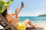 Fototapeta  - Attractive young woman sending online a love message through a selfie photo on the mobile phone while sitting on the beach during summer vacation