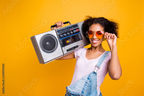 Zdjęcie XXL Portret fancy toothy dziewczyna z promiennym uśmiechem w okularach, trzymając boom box na ramieniu patrząc na kamery na białym tle na żółtym tle. Koncepcja hobby fanem muzyki