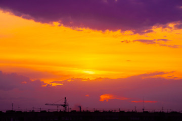 Wall Mural - Magenta sunrise above silhouette of city buildings with tower cranes. Unusual cityscape with beautiful dawn. Picturesque warm city landscape with bright sunny sky. Orange and purple cloudy layer sky.