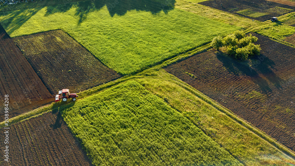 Aerial view from the drone, a bird's eye view of agricultural fields with a road through and a tractor on it in the spring evening at sunset - obrazy, fototapety, plakaty 
