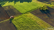 Aerial View From The Drone, A Bird's Eye View Of Agricultural Fields With A Road Through And A Tractor On It In The Spring Evening At Sunset