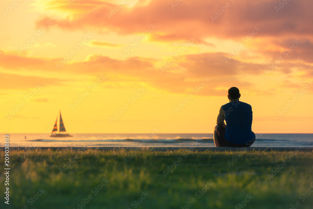 Man relaxing by the sea watching the colourful sunset and sail boats go ...