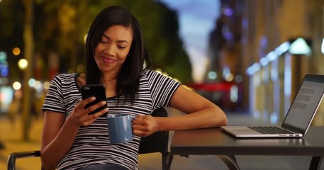 Wall Mural - Black woman writer on trip in Paris taking a break from work using cellphone, Cheerful African American female with her laptop and coffee texting on smartphone, 4k