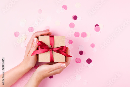 Woman's hands holding kraft gift box with red bow on pink background decorate...