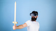 © be free - Gamer concept. Guy with head mounted display holds sword, play fighting game in VR. Hipster on serious face enjoy play game in virtual reality. Man with beard in VR glasses, light blue background