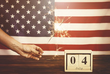 Retro Toned Cropped Shot, Woman Holding Burning Sparkler, Bengal Light On American Flag Background. 4th Of July, USA Independence Day Date On Wooden Cube Calendar. Vintage Filter, Copy Space, Close Up