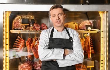 Male butcher standing near refrigerator with raw meat indoors