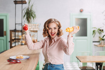 Pretty lady in shirt sitting with ketchup and mustard bottles in hands and joyfully looking in camera while spending time in cafe