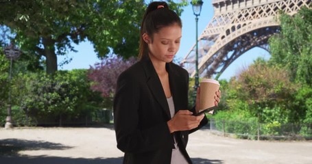 Wall Mural - Young professional woman on business trip in Paris sending text message under Eiffel Tower while drinking coffee, Millennial businesswoman using cell phone while meeting colleague in France, 4k