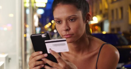 Wall Mural - Close up of young woman on city street shopping online with card and smartphone, Pretty Caucasian female standing on urban street at night paying for goods on Internet with mobile phone, 4k