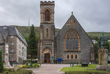 Fort William, Scotland - June 11, 2012: Frontal View On Duncansburgh Church Of Scotland, Macintosh Parish, Located On The Parade In Downtown Under Gray Sky With Hills In Back. Front, Two Statues.