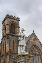 Fort William, Scotland - June 11, 2012: Closeup Of White Stone War Memorial Statue On The Parade Against Gray Sky. Shows Young Soldier In Kilt Contemplating, Duncansburgh Church Of Scotland In Back.