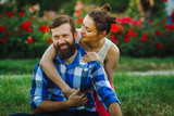 Fototapeta Uliczki - Close up portrait of attractive brunette woman and her handsome bearded boyfriend in park. Bush of roses flowers in the background