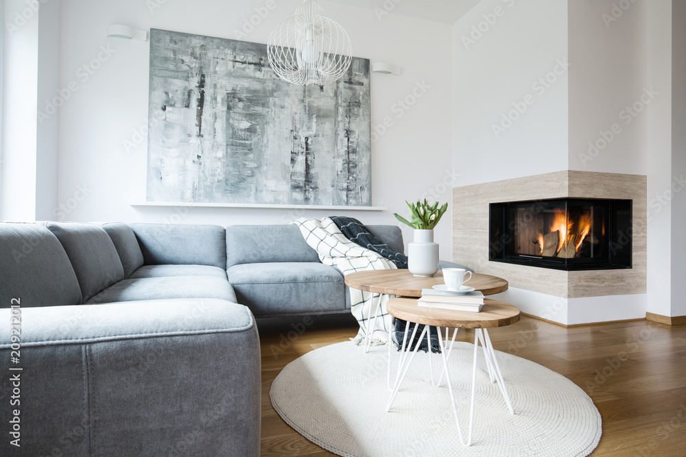 Grey corner settee with blankets standing in white Nordic living room interior with fresh tulips, books and tea cup on hairpin tables, abstract painting and fireplace - obrazy, fototapety, plakaty 