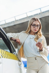 Wall Mural - beautiful blonde woman in eyeglasses leaning at taxi cab and using smartphone