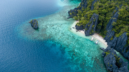 Aerial drone view of boats above a tropical coral reef and small sandy beach surrounded by huge cliffs (Secret Lagoon, Miniloc)