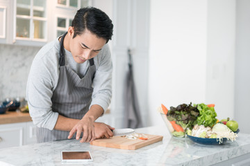 Asian confused man reading his tablet with a pensive thoughtful look while standing in his kitchen while cooking and preparing a meal