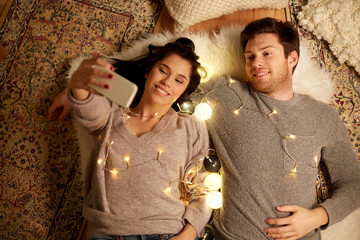 Sticker - leisure, hygge and people concept - happy couple with garland lying on floor at home and making selfie by smartphone