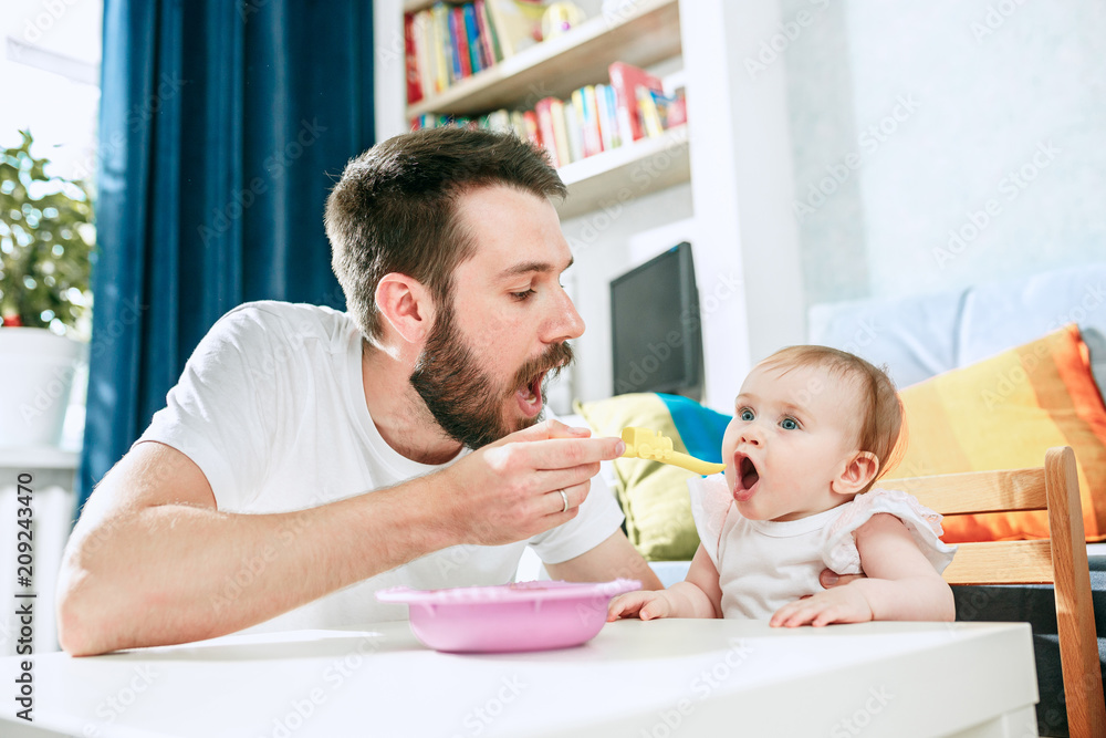 Good looking young man eating breakfast and feeding her baby girl at home - obrazy, fototapety, plakaty 