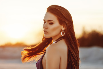 Outdoor close up portrait of young beautiful woman with long hair wearing dress, golden earrings, necklace, posing at the sunset