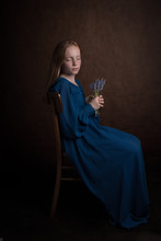 Classic Studio Portrait Of Girl In Blue Vintage Dress Sitting And Dreaming On Chair With Flowers
