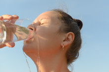 A Simple European Young Woman Is Drinking Water From A Glass Beaker Against A Blue Sky.