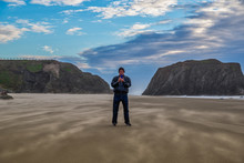 One Man Standing On A Beautiful Beach In Bandon Oregon Holding Up His Phone And Taking Pictures. Windy Conditions, Blue Sky With Nice Clouds