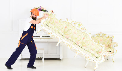 Wall Mural - Delivery service concept. Courier delivers furniture in case of move out, relocation. Loader moves sofa, couch. Man with beard, worker in overalls and helmet lifts up sofa, white background.