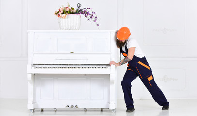 Wall Mural - Man with beard worker in helmet and overalls pushes, efforts to move piano, white background. Loader moves piano instrument. Courier delivers furniture, move out, relocation. Heavy loads concept.