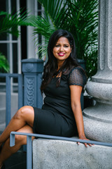 An image of a successful and attractive Indian Asian woman sitting on a bench while leaning on a concrete pillar in the park. She is wearing a very elegant,professional and classy black dress. 