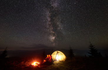 Young couple hikers man and woman having a rest near illuminated tent by burning campfire under starry sky with Milky way, enjoying quiet night camping in mountains. Tourism, beauty of nature concept.