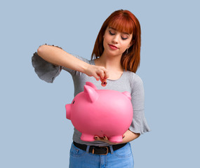 Young redhead girl holding a big piggybank on blue background