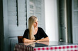 © Danon - Image of a young blonde woman with blue eyes sitting at a table of an old building. She is casually and comfortably dressed.