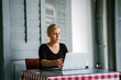 © Danon - A young and attractive blond woman with blue eyes works on her laptop in a cafe during the day.