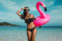 A Beautiful Sexy Amazing Young Woman On The Beach Sits On An Inflatable Pink Flamingo And Laughs, Has A Great Time, Tanned Perfect Body, Long Hair, Black Bikini, Fashion Accessories, Low Key Photo