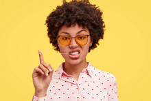 Brutal Attractive Dark Skinned Female With Afro Hairstyle, Stretches Chewing Gum And Looks With Displeased Expression, Wears Trendy Sunglasses, Isolated Over Yellow Background. Cool African Woman