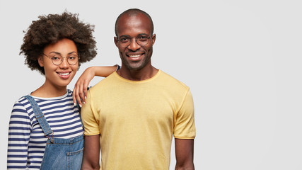 Wall Mural - Horizontal shot of pretty African American female leans at shoulder of his companion, have spare time, pose together over white wall with copy space for your advertisement. Happy dark skinned couple