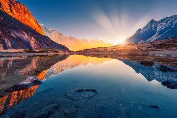 Wall Mural - Beautiful landscape with high mountains with illuminated peaks, stones in mountain lake, reflection, blue sky and yellow sunlight in sunrise. Nepal. Amazing scene with Himalayan mountains. Himalayas