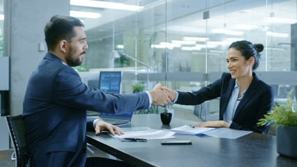 Businesswoman and Businessman Shake Hands. Draw up a Contract, Filling Papers in Conference Room. In the Background Modern Bright Office with Glass Walls.