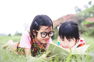 Wall Mural - Two little girls with glasses lying on the grass reading a book in garden at summer time