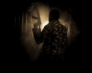 Silhouette of man with assault rifle ready to attack on dark toned foggy background or dangerous bandit in black wearing balaclava and holding gun in hand.