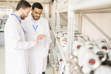 Wall Mural - Multi-ethnic team of quality control engineers wearing lab coats studying document while standing at spacious warehouse of pressure transducer factory and carrying out inspection