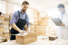 Confident Middle-aged Worker Wearing Checked Shirt And Jumpsuit Packing Manometers In Cardboard Box While His Colleague Filling In Receipt