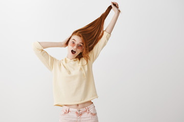 Wall Mural - Everyone is jealous of her beautiful hair. Portrait of playful expressive young redhead in fashionable clothes and pink glasses pulling hair up with hand and gasping from excitement over gray wall